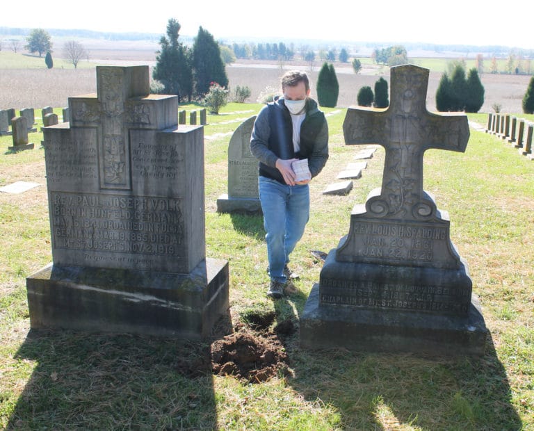 Edward Wilson prepares to place the stack of prayer cards in the ground next to the tombstone of Father Volk.
