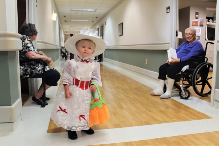 Ella, a miniature “Mary Poppins” and daughter of Anna Carman who works in Health Care in the Villa, poses before she makes her way down the hall. Looking on is Sister Sara Marie Gomez, right.