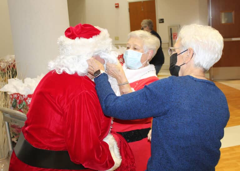 There are always new duties to being Santa’s wife. Here Mrs. Claus (Associate Elaine Wood) and Associate Betty Boren help Santa with his mask before entering Saint Joseph Villa.