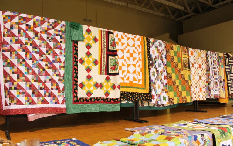 Multi-colored quilt tops adorn the stage in Maple Hall. Some of them will be hand quilted for the Mount Quilt Club drawings, others will be machine quilted for Quilt Bingo on Sept. 8.