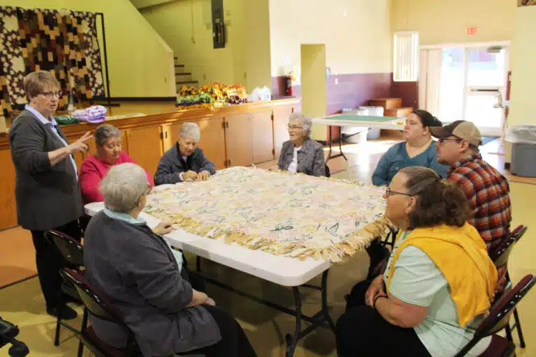 Sister Amelia Stenger, standing, who coordinated the blanket making, gives instruction to Ursuline Associate Amy Payne, right, on March 11, 2025. Amy and Tom Payne brought participants from Commonwealth Connections – which serves adults with disabilities – to help with the project. Two of the participants are seated next to Amy. Sisters working around the table, from left, are Sister Mary McDermott, Sister Rose Jean Powers, Sister Joan Riedley, and Sister Ann Patrice Cecil. Sister Amelia Stenger, standing, who coordinated the blanket making, gives instruction to Ursuline Associate Amy Payne, right, on March 11, 2025. Amy and Tom Payne brought participants from Commonwealth Connections – which serves adults with disabilities – to help with the project. Two of the participants are seated next to Amy. Sisters working around the table, from left, are Sister Mary McDermott, Sister Rose Jean Powers, Sister Joan Riedley, and Sister Ann Patrice Cecil.