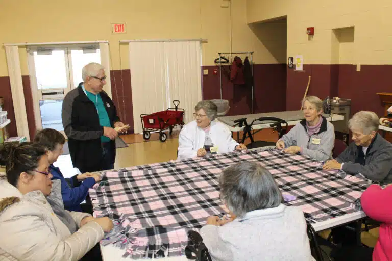 Ursuline Associate Tom Payne, standing, shares a laugh with Sister Margaret Ann Aull and other Sisters working on a blanket. Payne is program director for Commonwealth Connections, which serves adults with disabilities. Two participants from Commonwealth Connections are pictured at left, Cathy and Iris. They were especially fast at blanket making. Ursuline Associate Tom Payne, standing, shares a laugh with Sister Margaret Ann Aull and other Sisters working on a blanket. Payne is program director for Commonwealth Connections, which serves adults with disabilities. Two participants from Commonwealth Connections are pictured at left, Cathy and Iris. They were especially fast at blanket making.