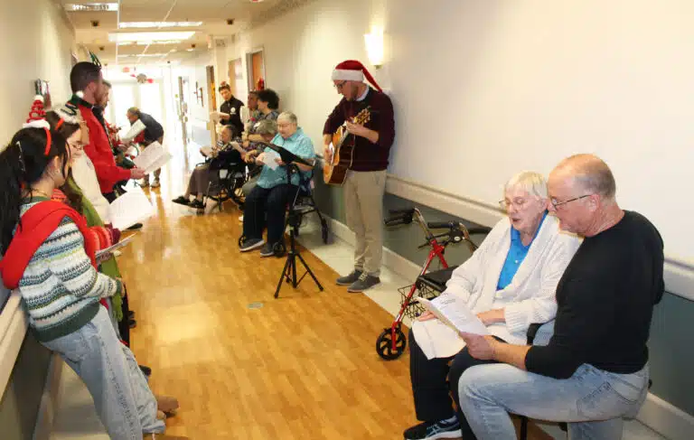 Bill Blincoe, right, director of Project and Risk Management for the diocese, takes a knee to share his song sheet with Sister Grace Simpson, as Danny May, director of Marriage and Family Life, plays the guitar and leads the caroling in the hallway of Saint Joseph Villa.