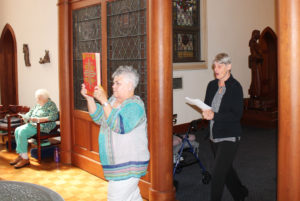 Associate Karen Wells, the first lector, carries in the lectionary, as the second lector, Cindy Bornander sings the entrance hymn “The Great Forerunner of the Morn.”