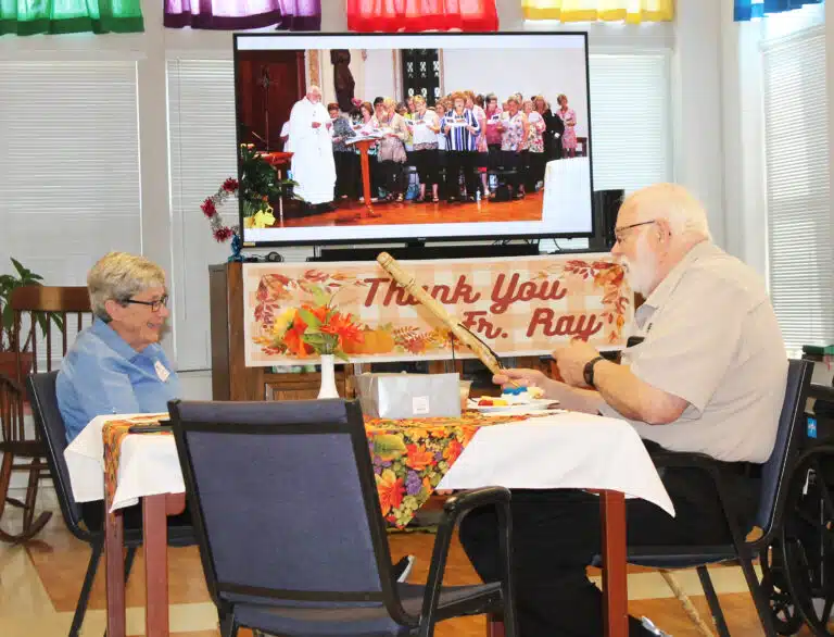 Father Ray shares a laugh with Sister Judith Nell Riney as a video of his time as Maple Mount chaplain plays in the background.
