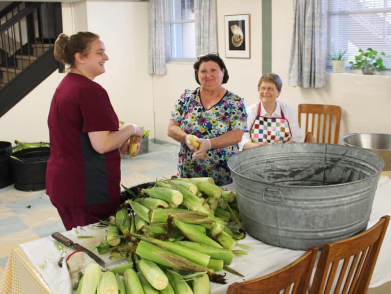 Food Service staff members Amanda Geary, left, and Charlotte Stelmach, center, enjoy shucking corn with Sister Amelia Stenger.