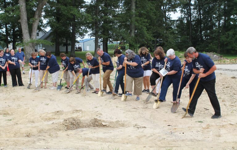 Teachers, the principal, school board members, two of Sister Anne Michelle’s sisters and Father Steve Hohman ceremonially dig into the hard ground to launch the construction project. The footer for the building has already been set behind them.