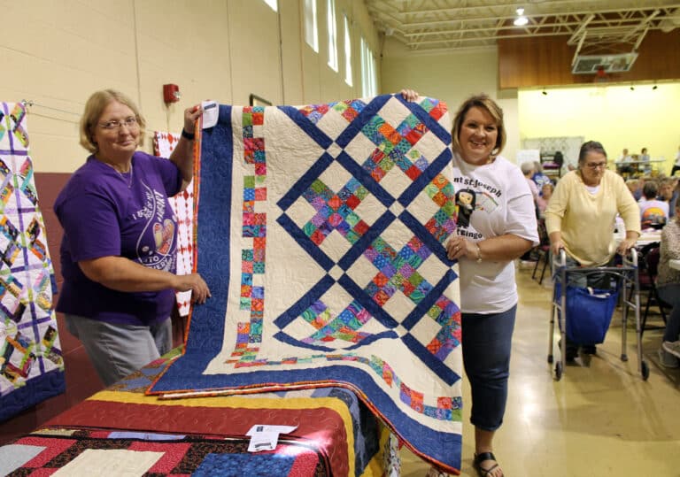 Cindy Clark of Alvaton, Ky., was wearing her Mount Saint Joseph Quilt Bingo shirt when she won a game and chose the large “Blue Beauty” quilt. Ann Jacobs, left, is helping her hold it.